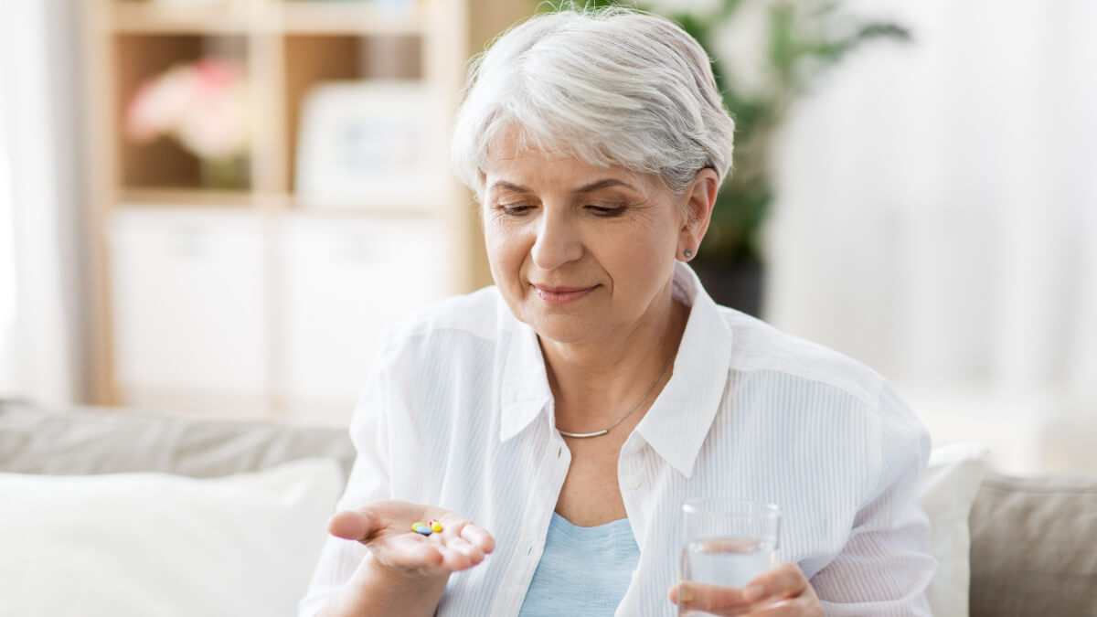 Mulher idosa segura suplementos em uma mão e um copo de água na outra. 
